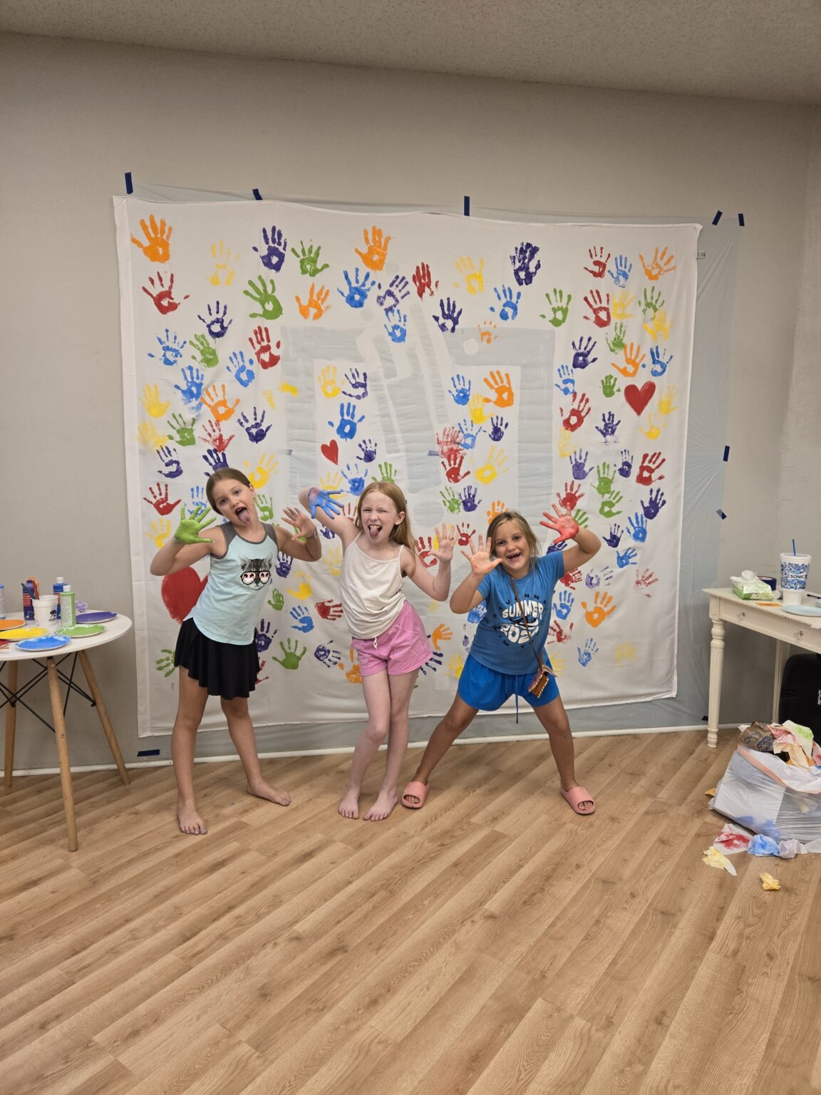 Three children with painted hands in front of a handprint banner during summer art class